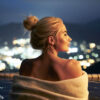 blonde woman, woman with blonde hair+hot spring, thermal bath+ bathing, taking a bath+bath towel, large towel used after bathing+night view, view of the city at night+ open-air bath, outdoor hot spring+soaking up to the shoulders, submerged up to the shoulders+hair tied up, hair pulled back+warm, cozy, comfortable+relaxed, unhurried, laid-back